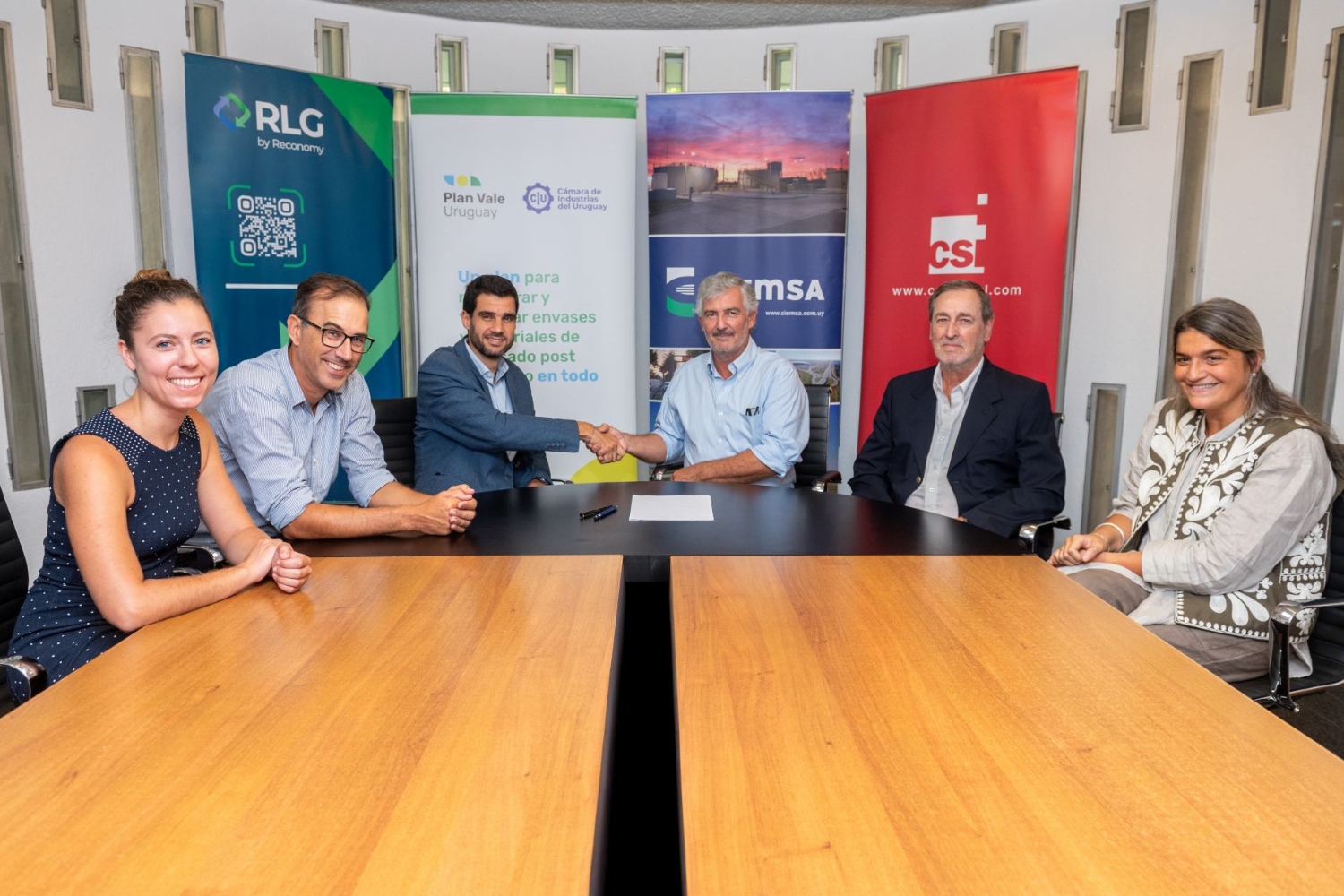 A group of six people sitting around a conference table, with two men in the center shaking hands, indicating a formal agreement or partnership. There are banners from various organizations in the background.