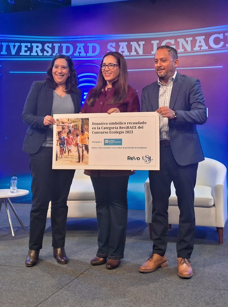 Three people standing together indoors holding a large symbolic donation check for the ReciRAEE category of the Ecologio 2023 contest at Universidad San Ignacio.