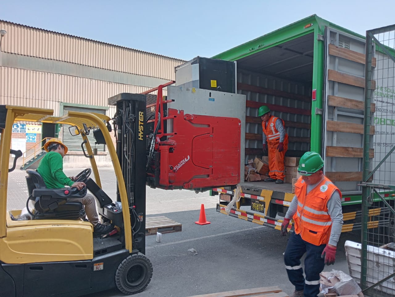 A forklift operator is unloading a container truck in an industrial setting. The operator is in a yellow forklift, wearing a green shirt and hat. Another worker stands inside the open truck, and a third person in an orange safety vest and green hard hat is standing nearby observing the process. The setting includes industrial buildings and safety cones.