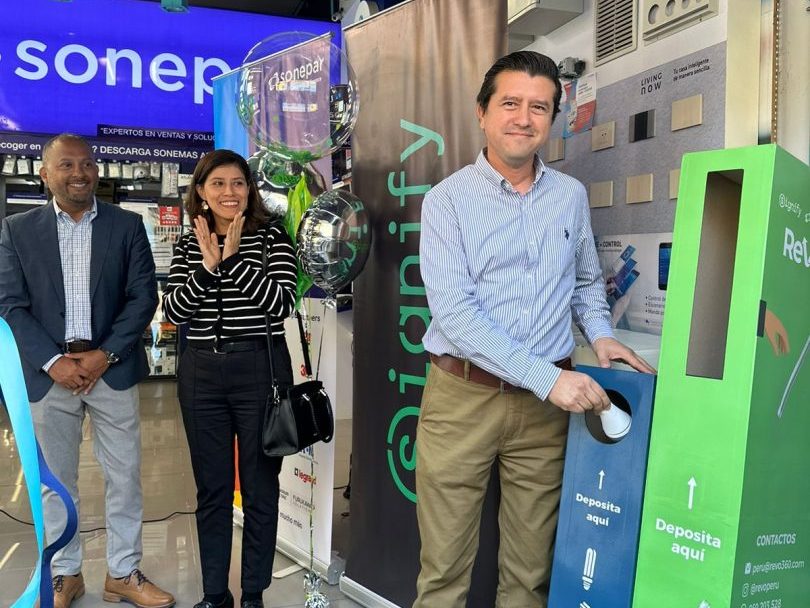Three people standing in a store with a blue and green recycling kiosk. One person is holding a light bulb above the kiosk slot, ready to deposit it. Store signage and product displays are visible in the background.