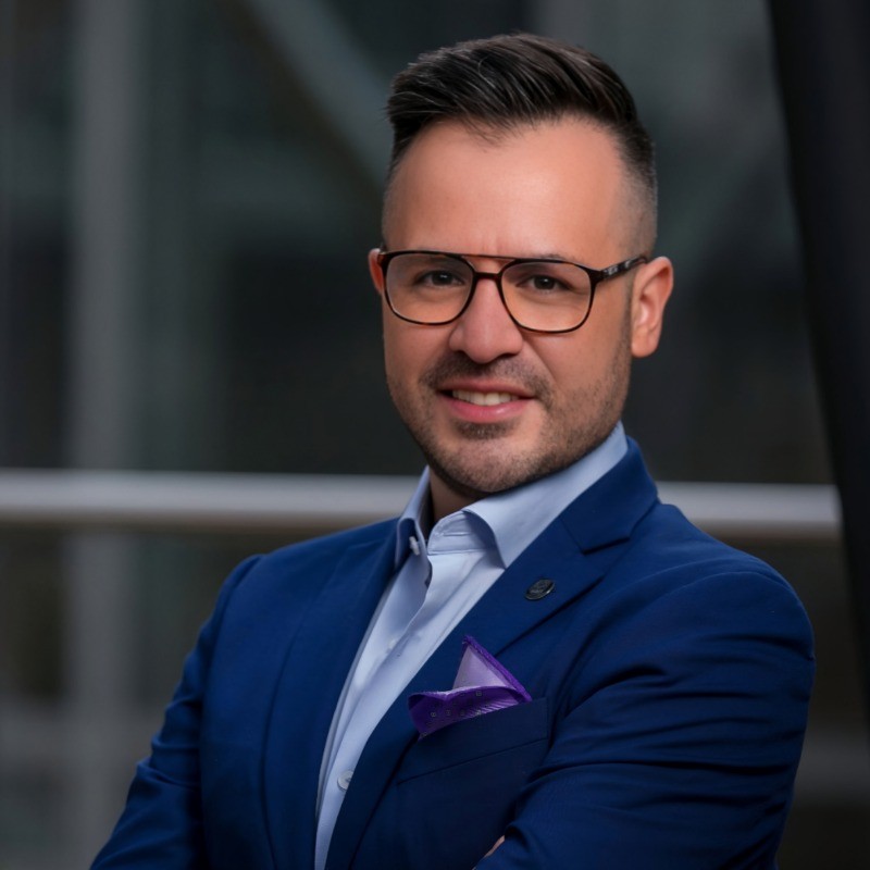 A man with short dark hair and glasses, wearing a blue suit and light blue shirt, standing with arms crossed and smiling.