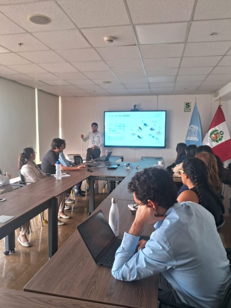 A man is giving a presentation in a conference room with a group of people seated around a long table. A presentation slide is displayed on a large screen at the front of the room. Two flags are visible in the background, one of which appears to be the flag of Peru. Attendees are using laptops and appear attentive to the presenter.