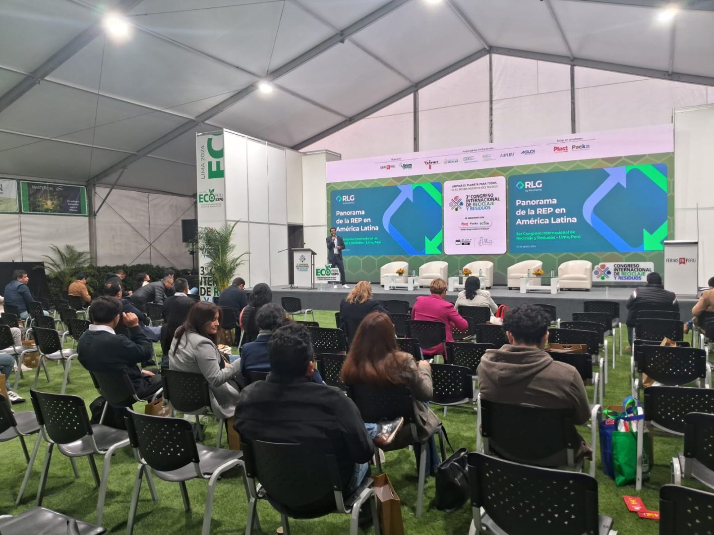 People seated in a conference room with green carpeting, listening to a speaker on stage in front of a large backdrop displaying 'Panorama de la REP en América Latina.'