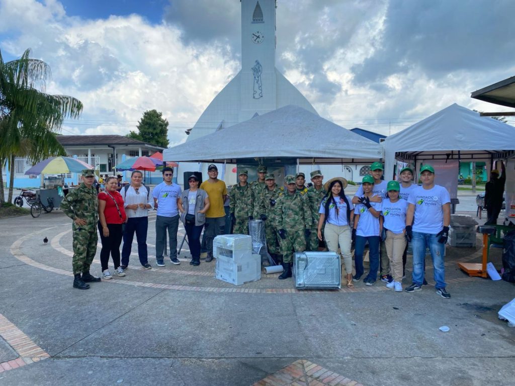 A group of people, including military personnel in uniform and civilians wearing white shirts with the text 'Fundación Verdes,' stand together outdoors in front of a tall building with a clock tower. They are posing for a photo beside white tents, with a few boxes on the ground.