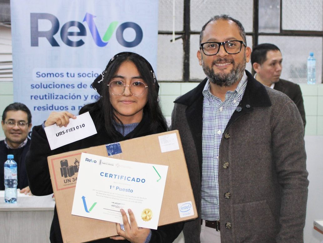 A young woman holding a certificate and a box poses with a man wearing glasses and a blazer in an indoor setting with a ReVO banner in the background.