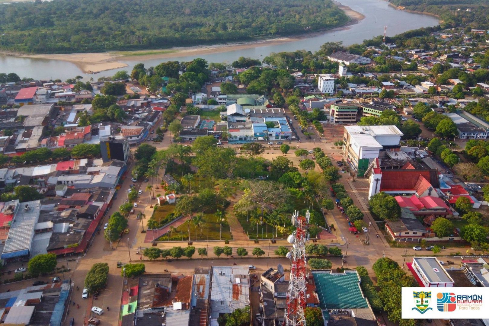 Aerial view of a town with a central park area surrounded by buildings, roads, and a river in the background. The landscape includes a mix of urban structures and greenery. A telecommunications tower is visible in the foreground. A logo with the text 'Ramón Guevara Para Todos' is in the bottom right corner.