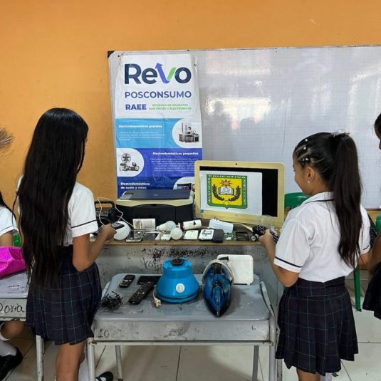 Young students in school uniforms stand at a table displaying various electronic devices as part of a recycling or educational exhibit on electronic waste, next to a poster labeled 'Re-Vo Posconsumo.'