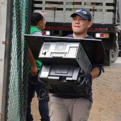 A man carrying a large electronic device, wearing a dark cap and gloves, standing near a truck and a chain-link fence.