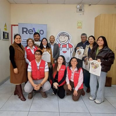 A group of people posing in a room, some wearing red vests with a logo, standing beside a cutout of a cartoon child with a large smile. Several individuals are holding tote bags with a similar cartoon design. A banner with 'ReYo' is visible in the background.
