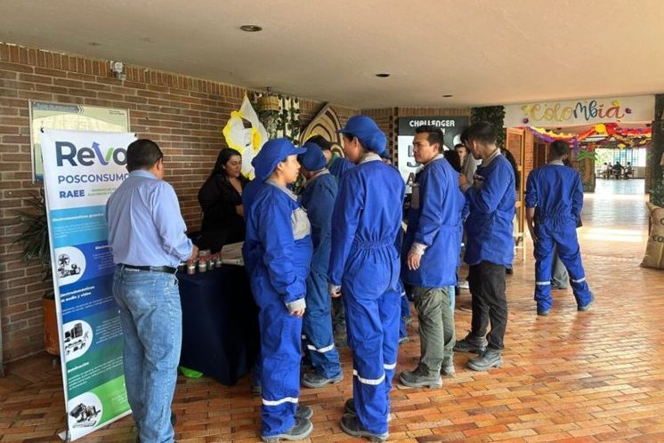 People in blue work uniforms gather around a table with a "Revo Posconsumo RAEE" banner, in a setting with brick walls and colorful decorations that include the word "Colombia."