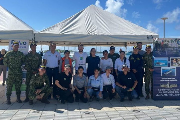 A group of people, including military personnel in camouflage uniforms and civilians, pose together under tents with banners at a naval event.