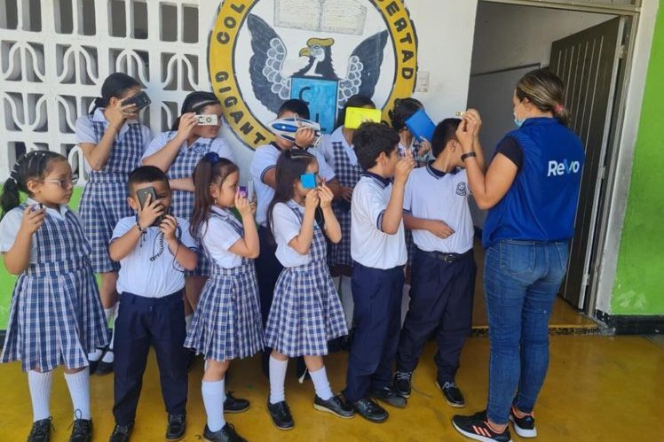 A group of school children in uniforms using VR headsets, guided by a woman in a blue vest, standing in front of a wall mural with the words 'Colegio Libertad Gigante'.