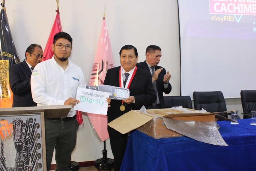 Two men standing in a room holding a sign and an LED light product; one man is wearing a white shirt with a company logo, the other is wearing a suit and medal, with flags and a projection screen in the background.