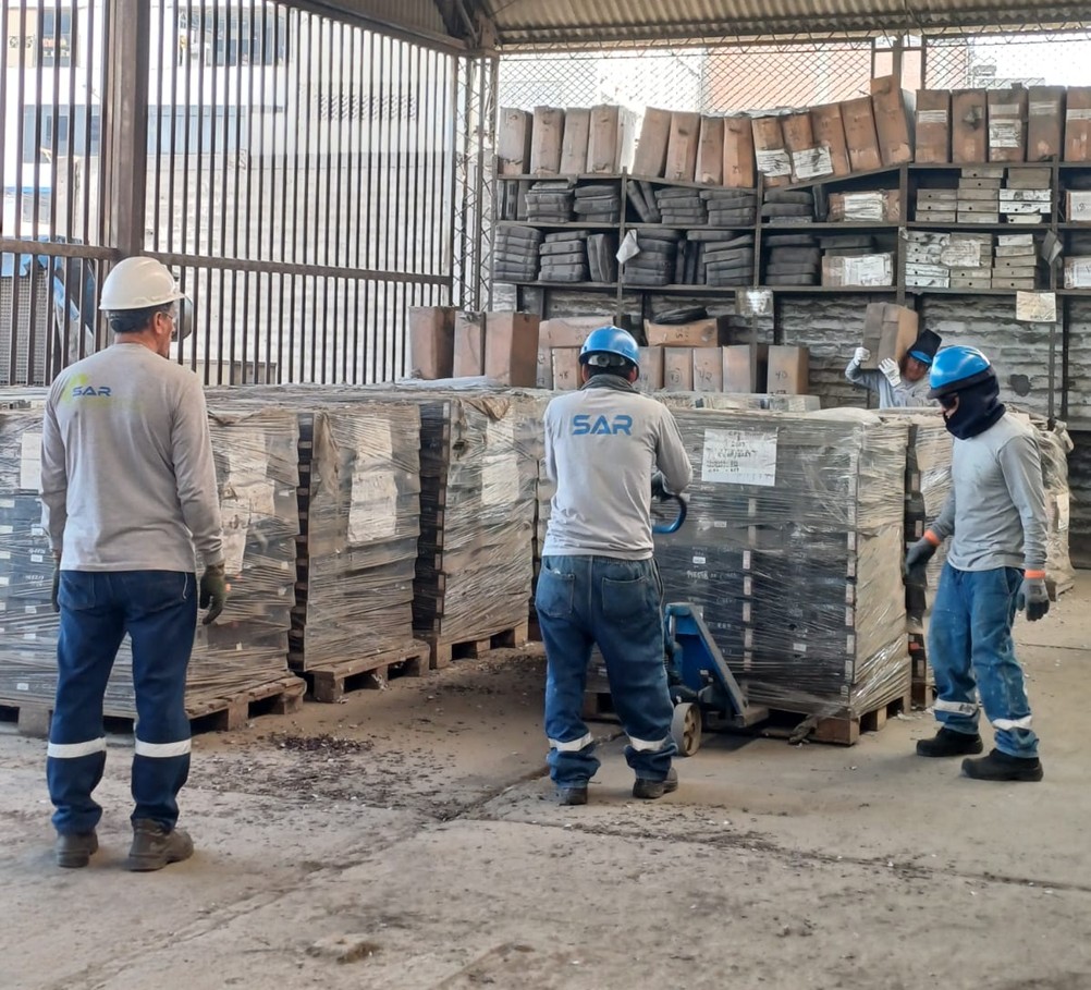 Workers in hard hats and protective clothing handle pallets of goods in an industrial warehouse with shelves stacked with boxes.